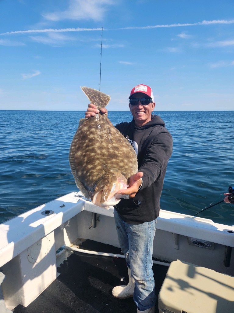 A man holding a large flounder while fishing on a boat, with water and blue sky in the background.
