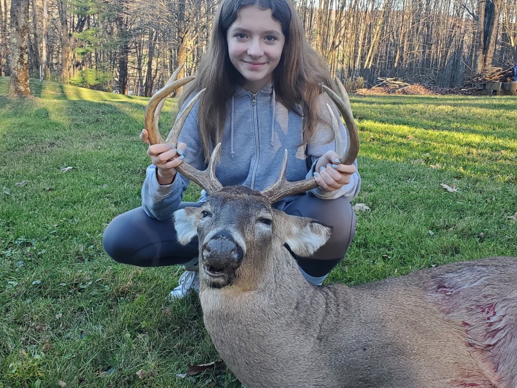 A young girl kneels beside a harvested deer on a grassy field, holding its antlers, with a wooded background.