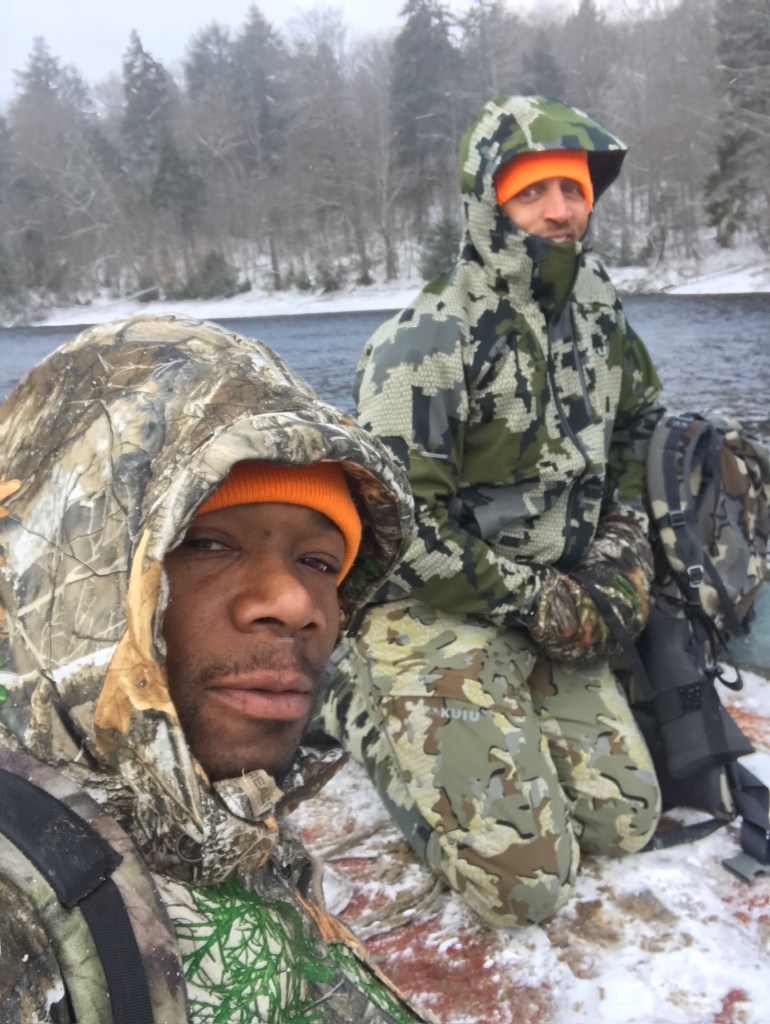 Two men wearing camouflage and hunting gear pose for a selfie in a snowy outdoor setting near a river.